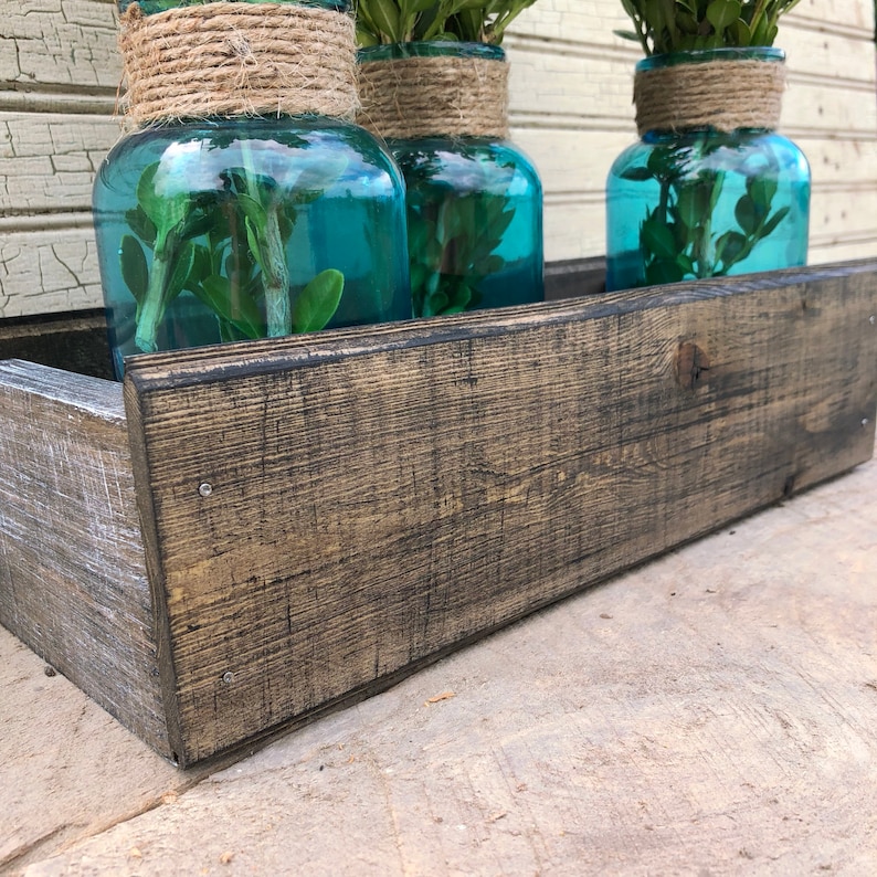 May include: Three teal blue glass jars with greenery inside, sitting in a rustic wooden planter box. The planter box is made of dark stained wood with a distressed finish.