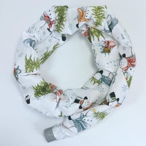 May include: A white fabric tube with a repeating pattern of snowmen, evergreen trees, and snowflakes. The snowmen wear hats and scarves in red, orange, and black. The tube has a gray plastic end.