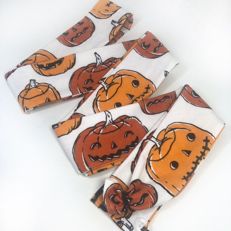 May include: White fabric headbands with a repeating pattern of orange and brown jack-o'-lanterns. The headbands are knotted and have a button closure. The Halloween-themed headbands are perfect for adding a festive touch to any outfit.