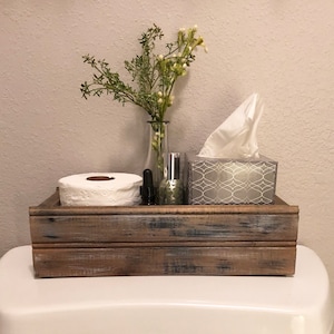 May include: A rustic wooden box with a distressed finish, containing a roll of toilet paper, a tissue box, a bottle of essential oil, and a small glass vase with greenery.