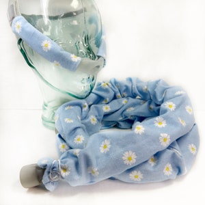 May include: Light blue fabric headgear with a white daisy pattern. The headgear is shown in two pieces, one wrapped around a clear glass head form and the other lying flat. The fabric is soft and the daisies have yellow centers.