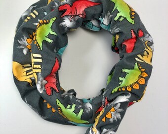 May include: Gray fabric neck gaiter with a dinosaur print. The dinosaurs are in various colors, including red, green, yellow, and blue. The gaiter has a black adjustable strap.