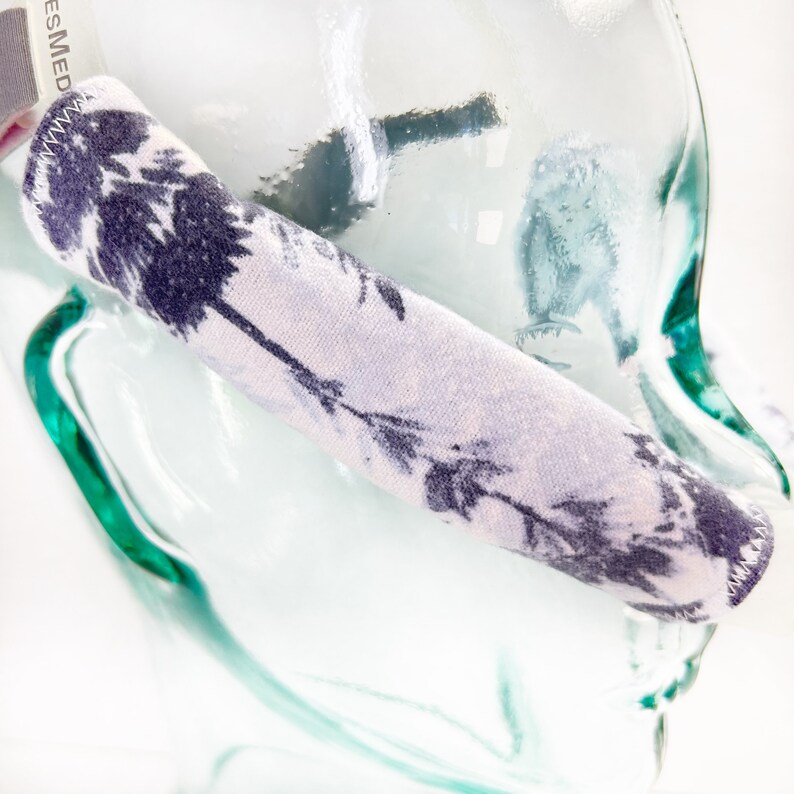 Pu&ograve; includere: Una fascia per capelli in tessuto con sfondo lavanda chiaro e una stampa floreale viola scuro di soffioni. La fascia presenta visibili cuciture a zigzag bianche lungo i bordi e un'etichetta parziale con la scritta ES MED.