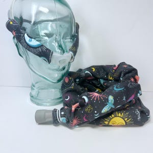 May include: A black fabric CPAP mask cover and tubing wrap with a celestial design featuring moons, stars, and suns in various colors. The mask cover has a moon and star design on the straps. The tubing wrap is rolled up.