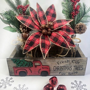 Puede incluir: Una caja de madera con un cami&oacute;n rojo pintado en la parte delantera, transportando un &aacute;rbol de Navidad verde. La caja tiene las palabras "&Aacute;rboles de Navidad reci&eacute;n cortados a la venta" pintadas en ella. La caja est&aacute; decorada con una poinsettia roja y negra a cuadros, ramas de pino y pi&ntilde;as.