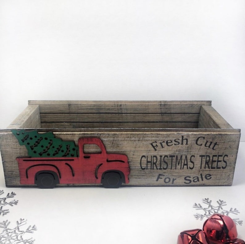 Puede incluir: Una caja de madera con un cami&oacute;n rojo que lleva un &aacute;rbol de Navidad verde. La caja tiene el texto "Fresh Cut Christmas Trees For Sale" impreso en ella.