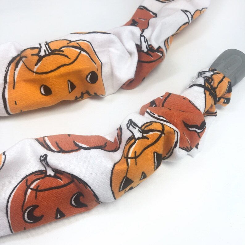 May include: White fabric with a repeating pattern of orange jack-o'-lantern faces. The fabric is wrapped around a gray object. The pumpkins have black eyes, noses, and mouths. The background is white.