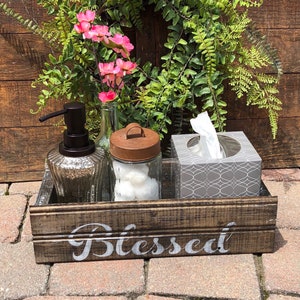 May include: A rustic wooden tray with the word "Blessed" in white lettering. The tray holds a glass soap dispenser, a jar with a rusty lid, a tissue box, and a small vase with pink flowers. A green wreath and wooden background complete the scene.