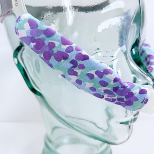 May include: A close-up of a clear glass head form with a floral patterned headband. The headband features a light blue background with purple and white floral designs. The headband is made of fabric.