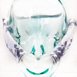 Pu&ograve; includere: Una coppia di copricinghia per maschera CPAP con un motivo a foresta grigio e bianco. Le coperture in tessuto sono progettate per avvolgere le cinghie laterali di una maschera, con cuciture a zigzag visibili lungo i bordi.