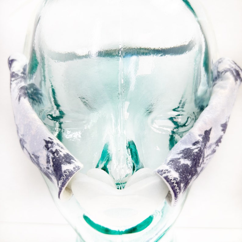 Pu&ograve; includere: Una coppia di copricinghia per maschera CPAP con un motivo a foresta grigio e bianco. Le coperture in tessuto sono progettate per avvolgere le cinghie laterali di una maschera, con cuciture a zigzag visibili lungo i bordi.