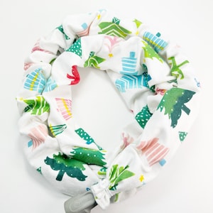 May include: A white fabric CPAP hose cover with a repeating pattern of colorful Christmas trees. The trees are in shades of green, pink, blue, and yellow. A gray ResMed connector is attached to the cover.