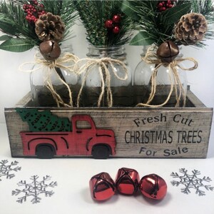 Puede incluir: Una caja de madera con un recorte de cami&oacute;n rojo y el texto "Fresh Cut Christmas Trees For Sale". Tres tarros de cristal con ramitas de vegetaci&oacute;n, bayas rojas y pi&ntilde;as est&aacute;n dispuestos en la caja. Los tarros est&aacute;n atados con cuerda y tienen campanas adjuntas.
