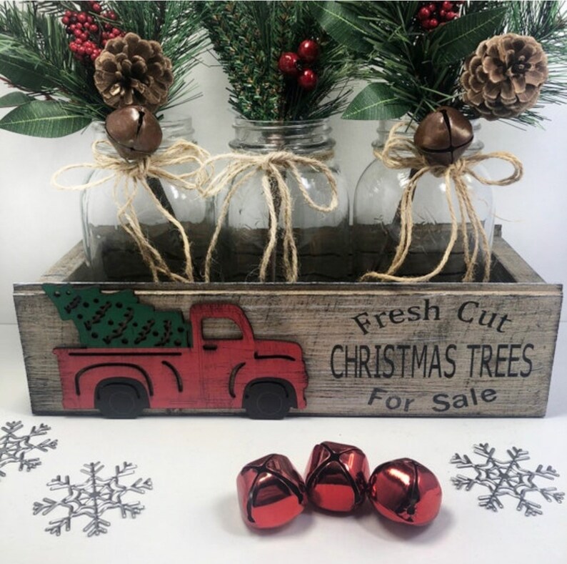 Puede incluir: Una caja de madera con un recorte de cami&oacute;n rojo y el texto "Fresh Cut Christmas Trees For Sale". Tres tarros de cristal con ramitas de vegetaci&oacute;n, bayas rojas y pi&ntilde;as est&aacute;n dispuestos en la caja. Los tarros est&aacute;n atados con cuerda y tienen campanas adjuntas.