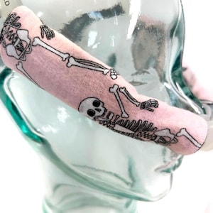 May include: A pink fabric sleeve with a black skeleton print. The sleeve is wrapped around a clear glass object. The skeleton design includes skulls, bones, and rib cages. The sleeve is made of a soft, possibly knitted material.