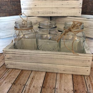 May include: Three clear glass mason jars with twine tied around the top, sitting in a white wooden box with a distressed finish.