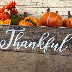 May include: A rustic wooden sign with the word "Thankful" painted in white. The sign is decorated with orange and brown fall-themed pumpkins.