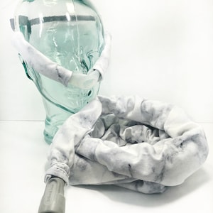 May include: A white and gray marble-patterned CPAP tube cover and headgear wrap. The headgear is on a glass head form. The tube cover is connected to a gray connector labeled "SlimLine".