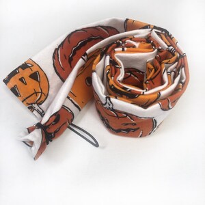 May include: Rolled-up Halloween-themed fabric featuring orange jack-o'-lanterns with black outlines on a white background. The fabric is rolled up, with a black elastic band visible at one end. The design is repeated across the fabric.