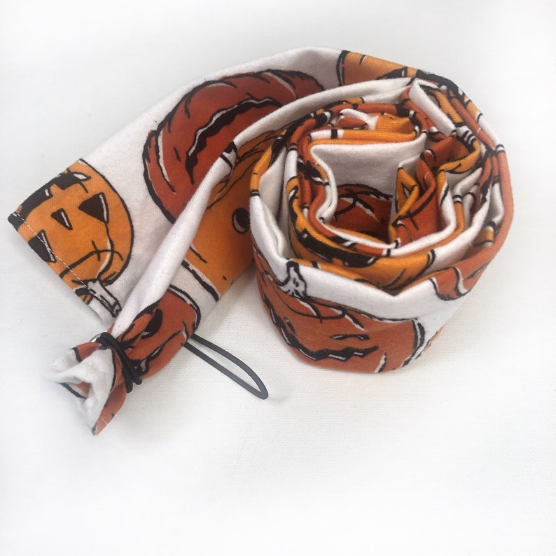 May include: Rolled-up Halloween-themed fabric featuring orange jack-o'-lanterns with black outlines on a white background. The fabric is rolled up, with a black elastic band visible at one end. The design is repeated across the fabric.