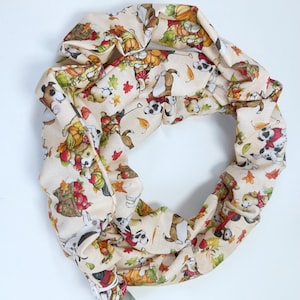 May include: Cream-colored infinity scarf with a repeating pattern of dogs, pumpkins, and autumn leaves. The dogs are depicted in various poses, some wearing scarves and carrying pumpkins. The scarf is made of a soft fabric and is suitable for fall.