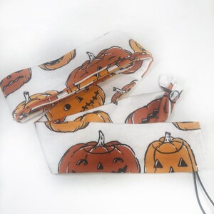 May include: White fabric with a repeating pattern of orange and brown jack-o'-lanterns. The fabric is folded and tied, with a small silver-colored ring. The design is perfect for Halloween decorations.