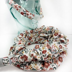 May include: A floral scrunchie and headband set. The white fabric is patterned with red, pink, and yellow flowers, along with green leaves. The scrunchie is bunched, and the headband is curved.