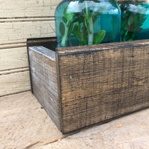 May include: A rustic wooden planter box with a dark brown stain and a distressed white finish. Two teal glass jars with greenery are sitting inside the planter box.