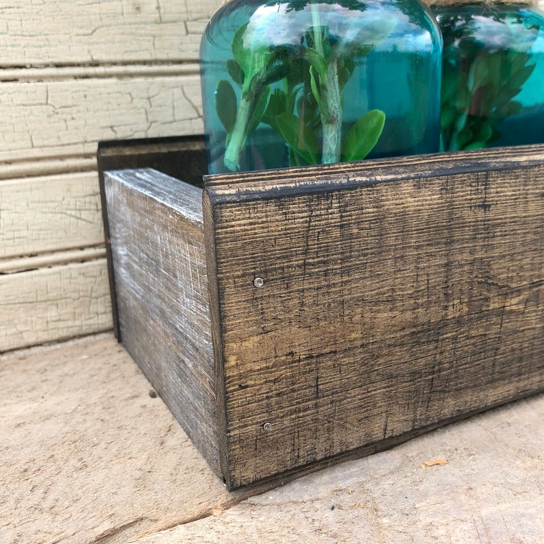 May include: A rustic wooden planter box with a dark brown stain and a distressed white finish. Two teal glass jars with greenery are sitting inside the planter box.