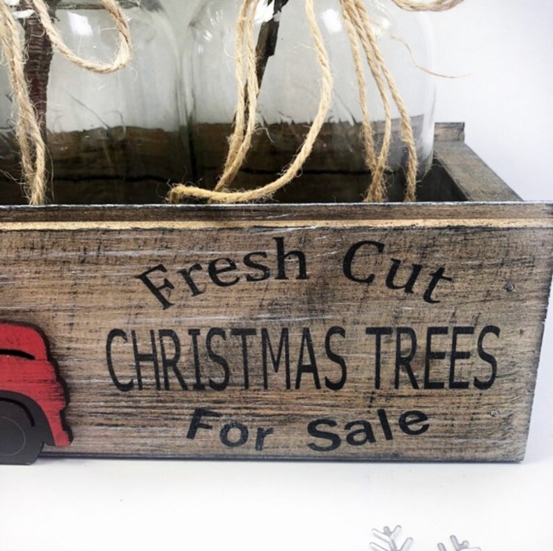 Puede incluir: Una caja de madera con el texto "Fresh Cut Christmas Trees For Sale" pintado en la parte delantera. La caja est&aacute; decorada con un cami&oacute;n rojo y tiene tres frascos de vidrio en su interior.