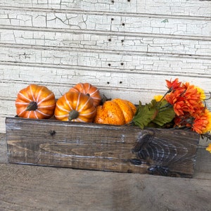 May include: A rustic wooden box filled with orange and yellow pumpkins and a bouquet of orange and yellow silk flowers. The box is sitting on a white painted wood surface.