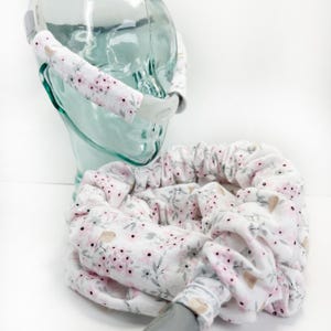 May include: A white and pink floral patterned fabric tube for a CPAP hose. The tube is wrapped around a clear glass head form. The fabric has a small floral pattern with pink flowers and green leaves.