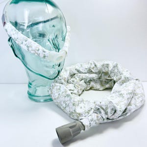 May include: A CPAP machine accessory set featuring a fabric cover for a nasal mask headgear and a matching fabric sleeve for a flexible air hose. Both items are made of white fabric with a repeating pattern of small green leaves and white flowers.