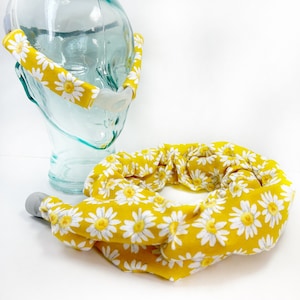 May include: A yellow fabric headband with a white daisy print. The headband is shown on a clear glass head form. The headband is also shown as a separate piece, rolled up in a circle.
