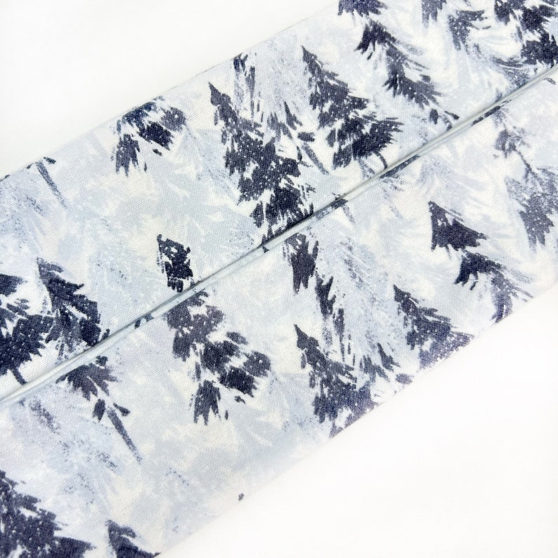 Pu&ograve; includere: Una striscia di tessuto con un motivo ripetuto di alberi sempreverdi blu navy scuro su uno sfondo bianco e grigio chiaro. Il tessuto presenta una trama visibile ed &egrave; piegato diagonalmente attraverso l'inquadratura.