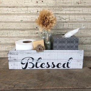 May include: A white wooden box with the word "Blessed" painted on the front. The box is filled with bathroom essentials, including a roll of toilet paper, a bar of soap, a bottle of hand sanitizer, and a box of tissues.