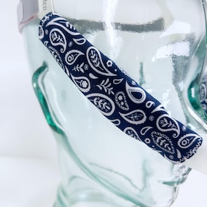 May include: A navy blue and white paisley pattern CPAP mask strap cover. The cover is wrapped around the strap of a clear glass CPAP mask. The paisley design features teardrop shapes and floral elements.
