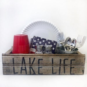 May include: A wooden crate with the words "Lake Life" painted on the front. The crate is filled with red plastic cups, white paper plates, blue and white napkins, silver utensils, and small glass salt and pepper shakers.