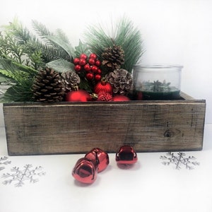 Puede incluir: Una caja de madera r&uacute;stica llena de adornos navide&ntilde;os, incluyendo ramas de pino, adornos rojos, pi&ntilde;as y un portavelas de vidrio con una vela verde. La caja est&aacute; sobre una superficie blanca con cascabeles rojos esparcidos alrededor.