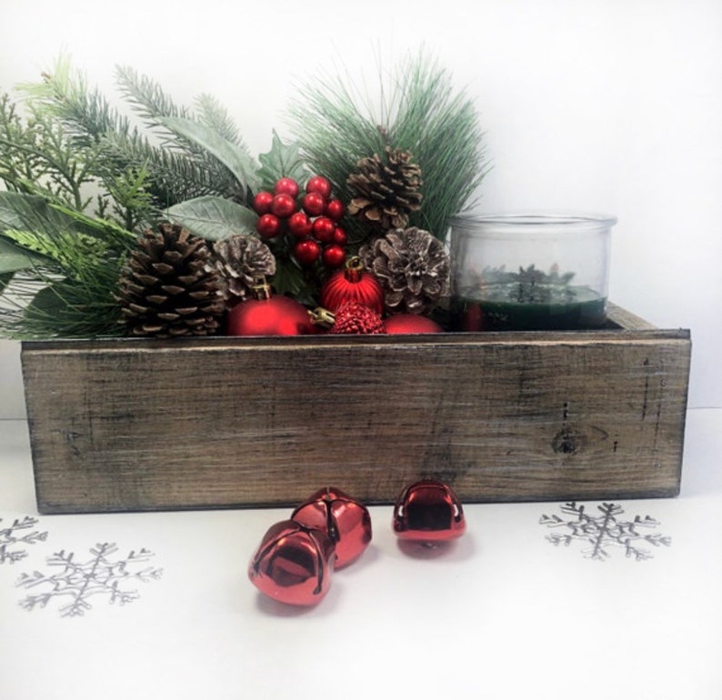 Puede incluir: Una caja de madera r&uacute;stica llena de adornos navide&ntilde;os, incluyendo ramas de pino, adornos rojos, pi&ntilde;as y un portavelas de vidrio con una vela verde. La caja est&aacute; sobre una superficie blanca con cascabeles rojos esparcidos alrededor.