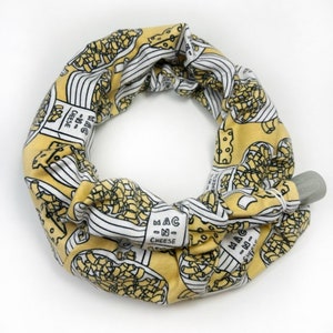 May include: A yellow and white fabric neck warmer with a repeating pattern of macaroni and cheese boxes. The boxes have the text "Mac 'n Cheese" on them.