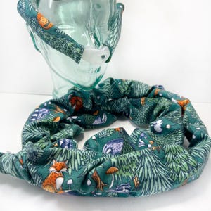 May include: A green fabric sleep mask with a woodland print featuring foxes, mushrooms, and trees. The mask is attached to a long green fabric tube with the same print.