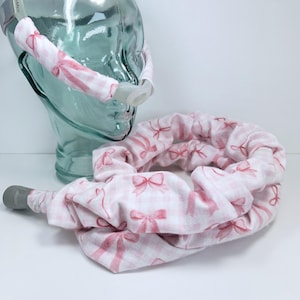 May include: A CPAP mask with a pink bow pattern on a white background. The mask is attached to a headgear with matching fabric. The design features a repeating pattern of pink bows on a white checkered background.