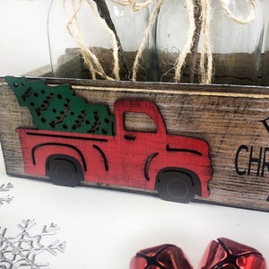 Puede incluir: Una caja de madera con un cami&oacute;n rojo recortado que lleva un &aacute;rbol de Navidad verde. La caja tiene las palabras "Primera Navidad" escritas en ella.
