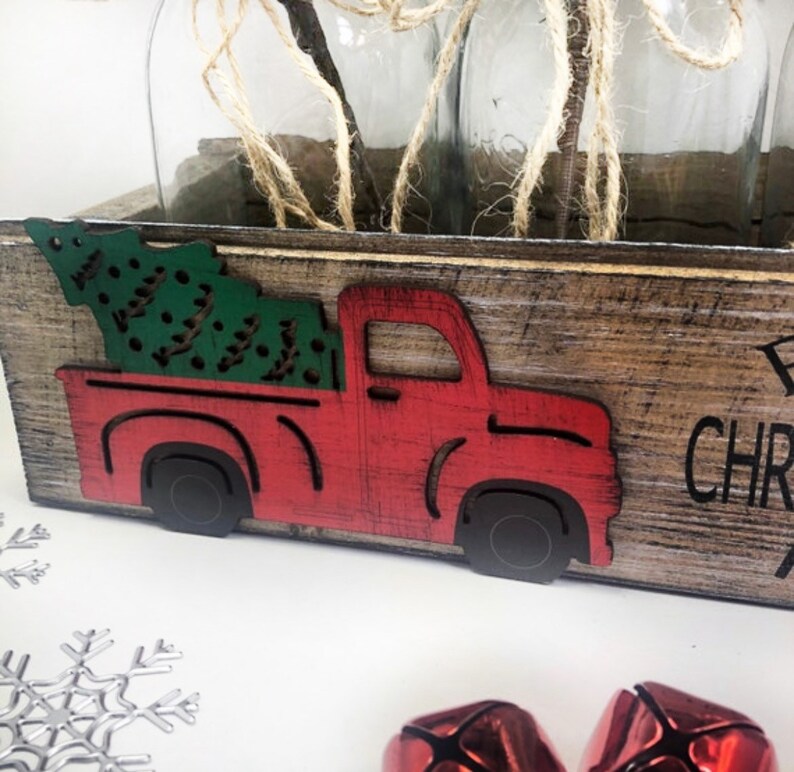 Puede incluir: Una caja de madera con un cami&oacute;n rojo recortado que lleva un &aacute;rbol de Navidad verde. La caja tiene las palabras "Primera Navidad" escritas en ella.