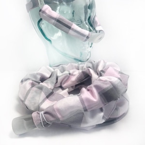 May include: A pink and gray plaid fabric cover for a CPAP hose. The cover is made of soft, stretchy fabric and is designed to fit snugly around the hose. The cover is shown on a clear plastic head model.