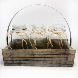 May include: Three clear glass jars with twine tied around the top and a bow, sitting in a rustic wooden box with a metal handle.