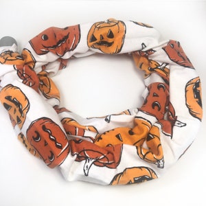 May include: A white fabric Halloween scarf with a repeating pattern of orange jack-o'-lantern faces. The scarf is looped into a circle, with a gray clasp at one end. The pumpkins have black details.