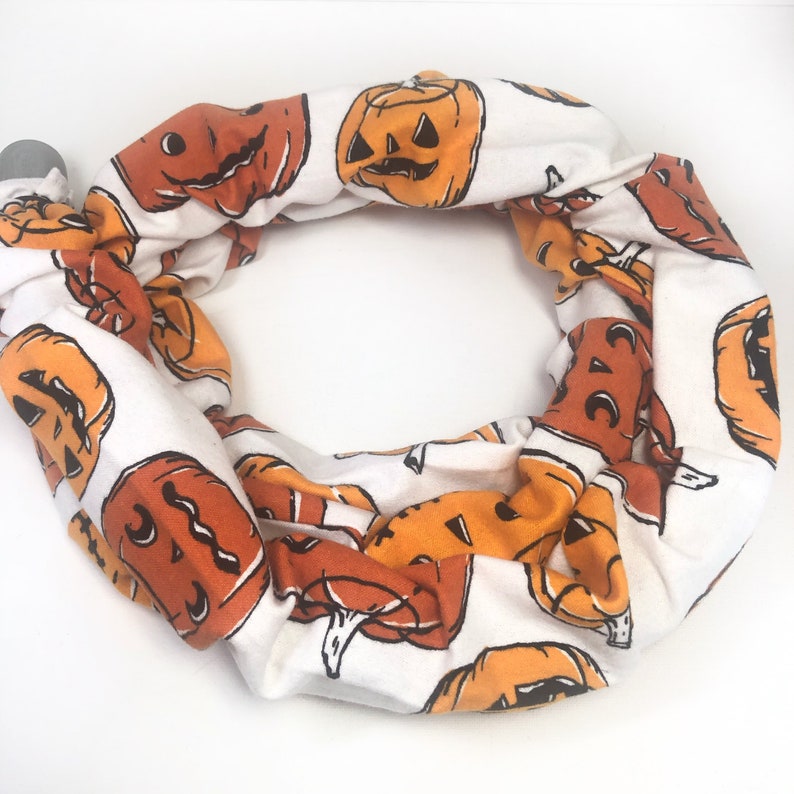 May include: A white fabric Halloween scarf with a repeating pattern of orange jack-o'-lantern faces. The scarf is looped into a circle, with a gray clasp at one end. The pumpkins have black details.