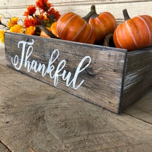 May include: A rustic wooden box with the word "Thankful" painted in white on the front. The box is filled with orange and yellow fall-themed decorations, including pumpkins and flowers.